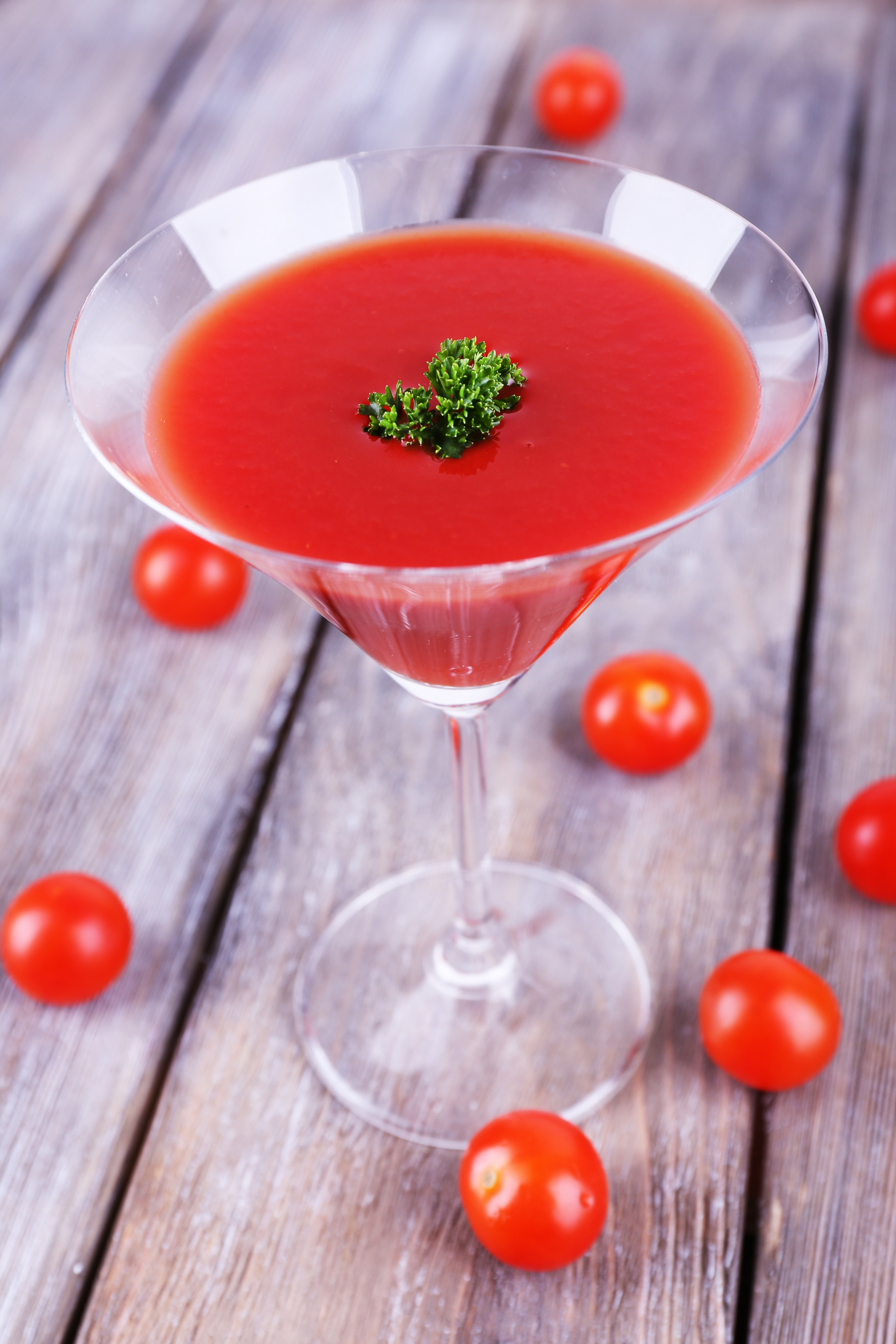 Red and bright tomato shrub cocktail on a wooden table with a sprinkle of parsley on top and tomatoes lying around. 