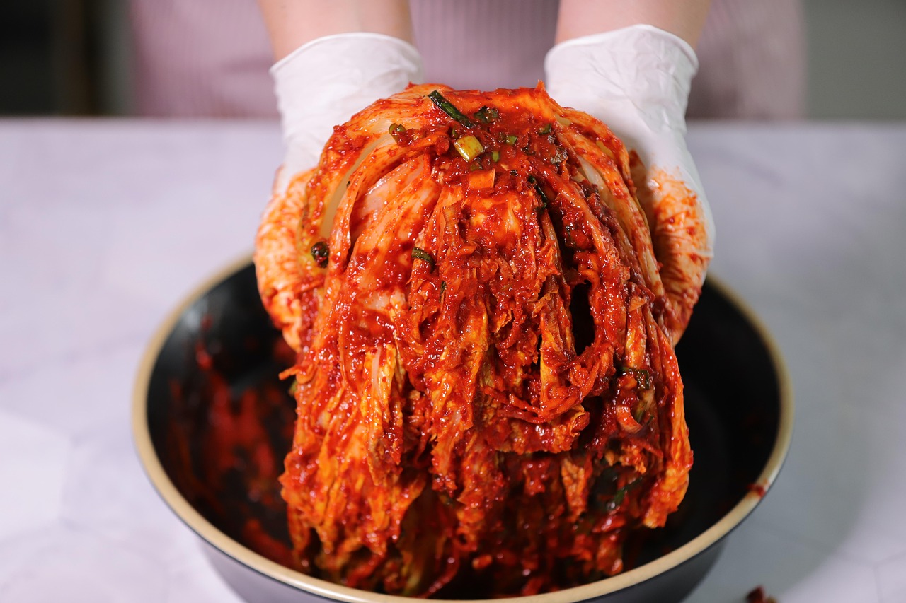 Gloved hands holding up a large portion of kimchi: fermented napa cabbage coated in a red chili pepper paste, over a bowl during the preparation process.