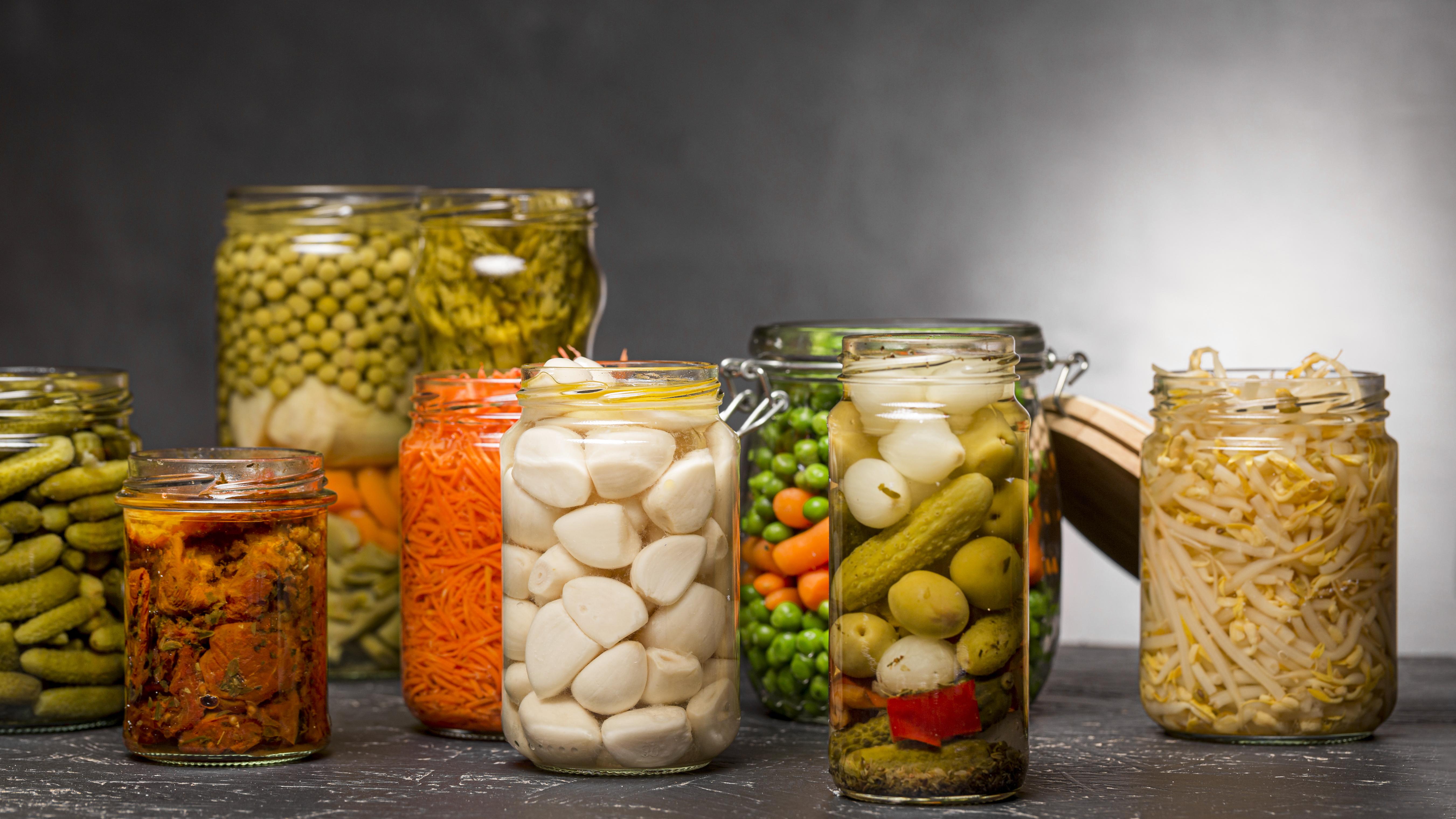 A colorful assortment of pickled vegetables in clear glass jars, including garlic cloves, carrots, gherkins, olives, peas, and bean sprouts, arranged on a dark surface with a gray background.