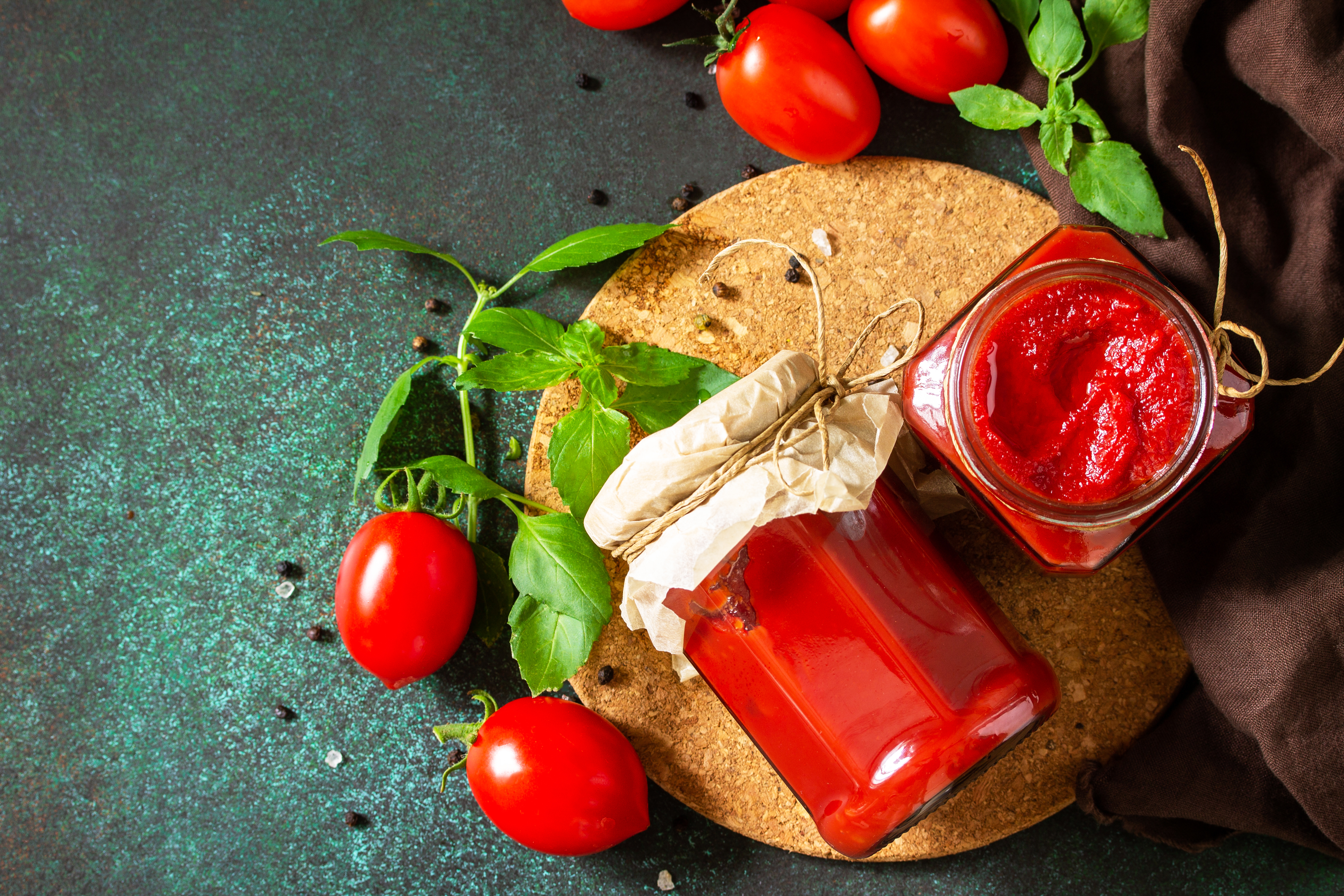 Jar with bright red tomato jam and a beige cover laying on top of a cork plate decorated with basil and cherry tomatoes. 