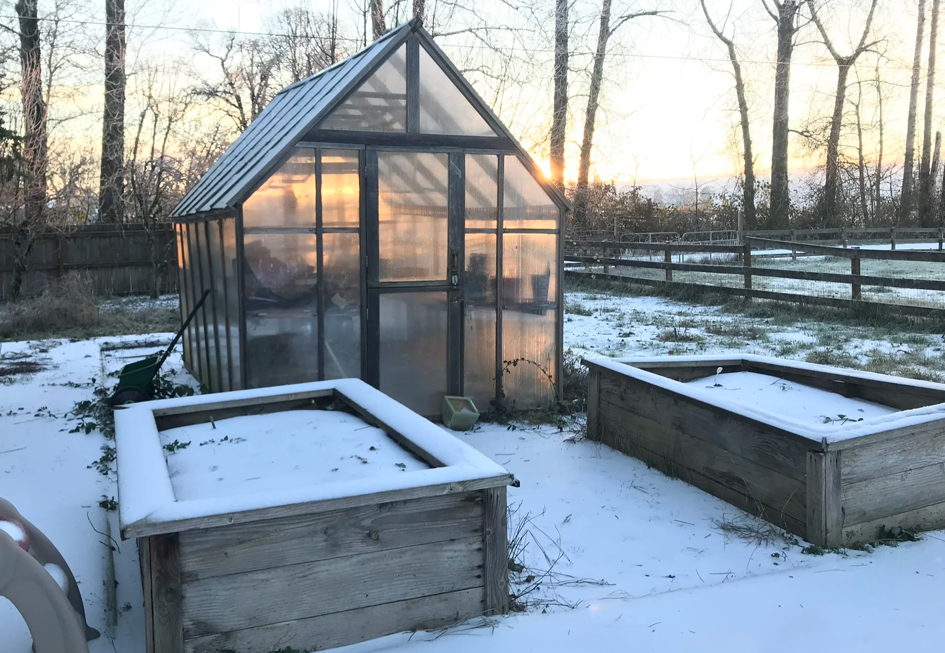 A glass greenhouse and raised garden beds covered in snow with a sunset on the horizon